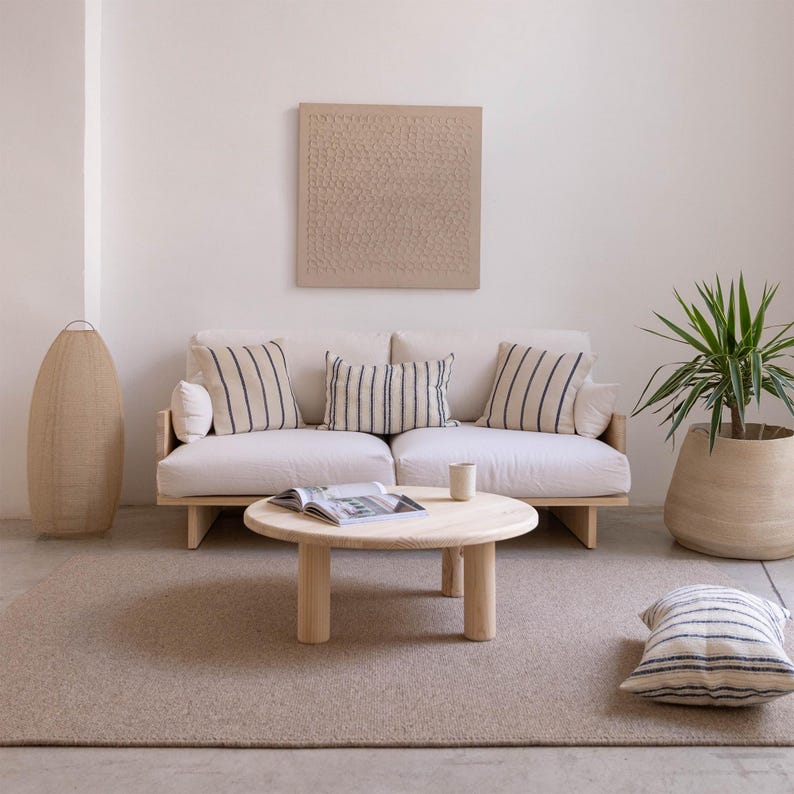 May include: A modern living room setup featuring a light-colored sofa with striped pillows, a round wooden coffee table, and a woven floor lamp. A potted plant and a textured wall art piece complete the minimalist design.