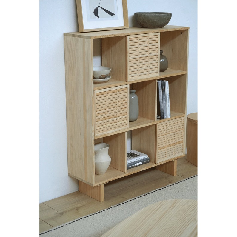Op de afbeelding: Een boekenkast van licht hout met lamellen deuren en open planken. De planken zijn versierd met vazen, kommen en boeken. De boekenkast staat op een lichte houten vloer.