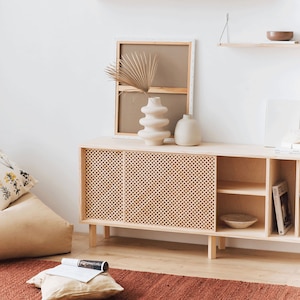 May include: A light wood cabinet with two sliding doors featuring a woven rattan pattern. The cabinet has two open shelves and is decorated with a dried palm leaf, a white ceramic vase, and a beige ceramic vase.