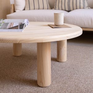 May include: A round, light-colored wooden coffee table with four cylindrical legs. A magazine and a ceramic mug rest on the table's surface. The table is set on a neutral-toned carpet, with a sofa in the background.