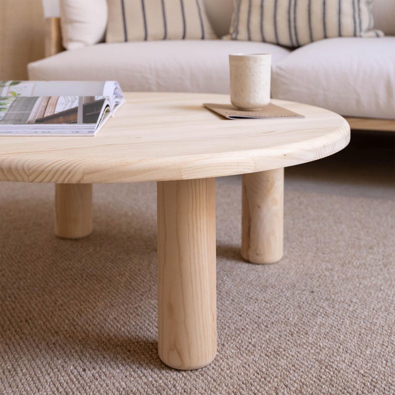 May include: A round, light-colored wooden coffee table with four cylindrical legs. A magazine and a ceramic mug rest on the table's surface. The table is set on a neutral-toned carpet, with a sofa in the background.