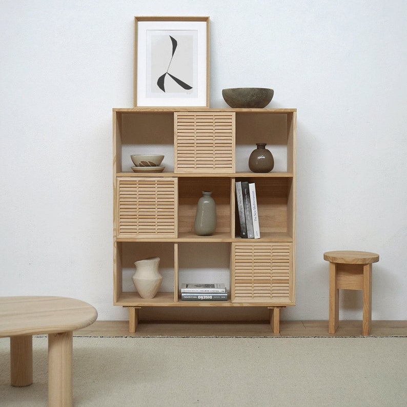 Op de afbeelding: Een houten boekenkast met lamellen deuren en open planken. De planken zijn versierd met boeken, vazen, kommen en een ingelijst drukwerk. De boekenkast staat op een lichtbruin tapijt.