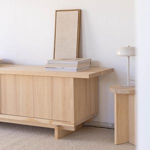 May include: A light wood cabinet with three doors and a light wood top. A stack of magazines sits on top of the cabinet. A white lamp with a round shade sits on a small light wood side table.