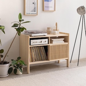 May include: A light wood record player stand with a record player, amplifier, and vinyl records. The stand has a slatted door and two shelves. The stand is in a room with a white wall and a floor lamp.