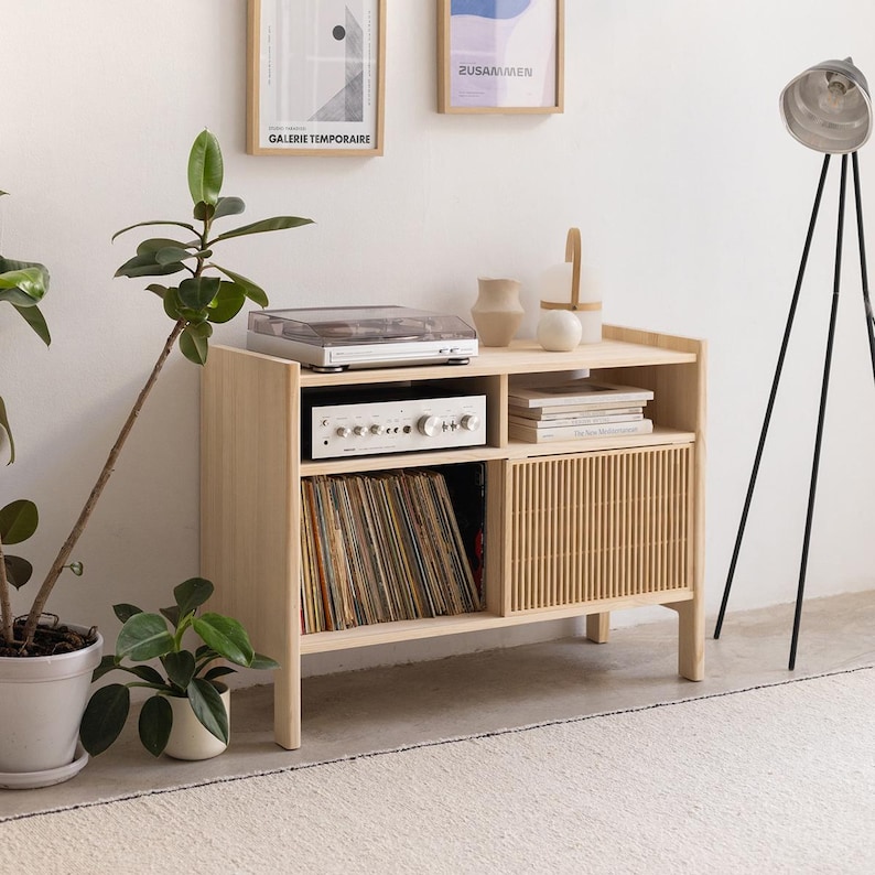 May include: A light wood record player stand with a record player, amplifier, and vinyl records. The stand has a slatted door and two shelves. The stand is in a room with a white wall and a floor lamp.
