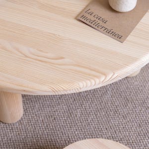 May include: A light-colored, round wooden coffee table with three short, cylindrical legs. The table's surface displays the natural wood grain. A small, rectangular card with the text "La casa mediterr&aacute;nea" rests on the table.