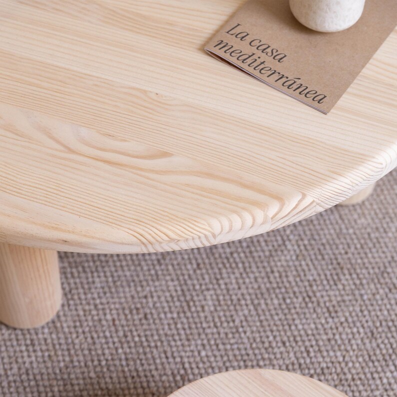 May include: A light-colored, round wooden coffee table with three short, cylindrical legs. The table's surface displays the natural wood grain. A small, rectangular card with the text "La casa mediterr&aacute;nea" rests on the table.
