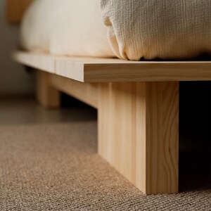Puede incluir: Una estructura de cama de madera de color claro con un dise&ntilde;o minimalista. La cama presenta una plataforma rectangular gruesa sostenida por patas de madera verticales y resistentes. Un coj&iacute;n de tono neutro descansa sobre la plataforma. La cama est&aacute; sobre una alfombra texturizada de color marr&oacute;n claro.