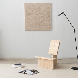 May include: A light wood chair with a simple design, featuring a square back and a wide seat. The chair is placed on a white rug in a room with a white wall and a textured beige artwork hanging above it.