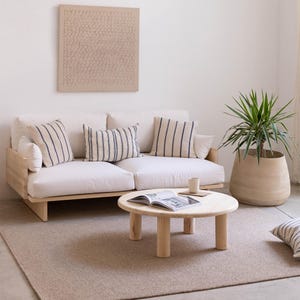 May include: A modern living room setup featuring a light wood sofa with cream cushions and striped pillows. A round wooden coffee table holds a book and a mug. A large potted plant and a textured wall art piece complete the space.