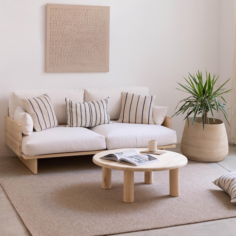 May include: A modern living room setup featuring a light wood sofa with cream cushions and striped pillows. A round wooden coffee table holds a book and a mug. A large potted plant and a textured wall art piece complete the space.