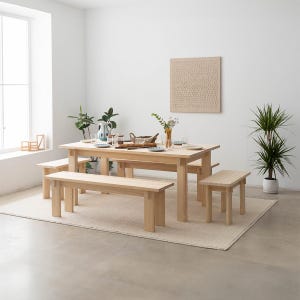 May include: A light wood dining table with four matching benches. The table is set with plates, glasses, and a bowl of fruit. There are two plants in the background, one on the left and one on the right.
