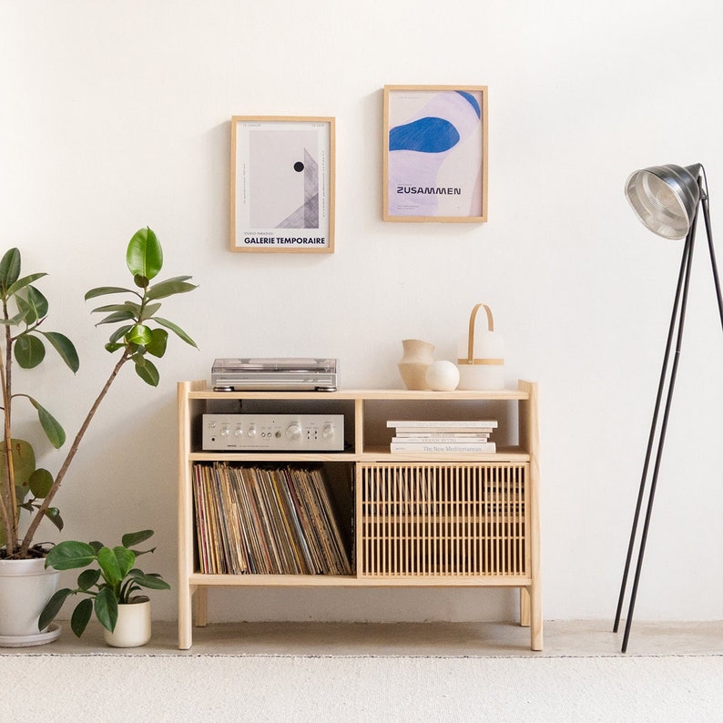 May include: A light wood cabinet with two shelves and a cabinet door. The cabinet is filled with vinyl records and has a stereo receiver on the top shelf. There are two framed prints on the wall behind the cabinet, one with the text "Galerie Temporaire" and the other with the text "Zusammen".