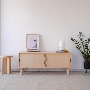 May include: A light wood cabinet with two doors that have a wavy design. The cabinet is sitting on a light beige carpet. There is a framed print on the wall above the cabinet, a small ceramic jar on top of the cabinet, and a white ceramic lamp with a stack of books on top of the cabinet. A potted plant is on the right side of the image.