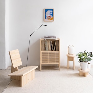May include: A light wood record player cabinet with a record player on top and shelves for vinyl records. The cabinet has a slatted door. A light wood chair, a small light wood table with a lamp, and a potted plant are also in the image.