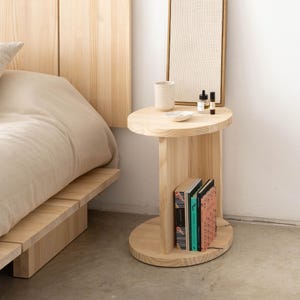 Puede incluir: Una mesita de noche de madera clara con una parte superior y una base redondas, y un soporte vertical con un estante. La mesa contiene una peque&ntilde;a taza de cer&aacute;mica, un plato y dos botellas. Dos libros est&aacute;n en el estante. Un marco de cama y un cabecero est&aacute;n en el fondo.