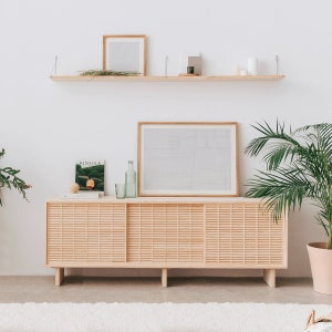 May include: A light wood cabinet with three doors and a slatted design. The cabinet is sitting on a white rug in front of a white wall. There is a shelf above the cabinet with a framed picture, a plant, and a few decorative items.