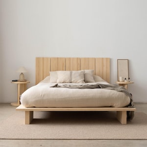 Puede incluir: Una cama de madera minimalista con cabecero claro y perfil bajo. La cama est&aacute; hecha con ropa de cama y almohadas de tonos neutros. Una peque&ntilde;a mesita auxiliar con una l&aacute;mpara y un espejo decorativo completan la escena.