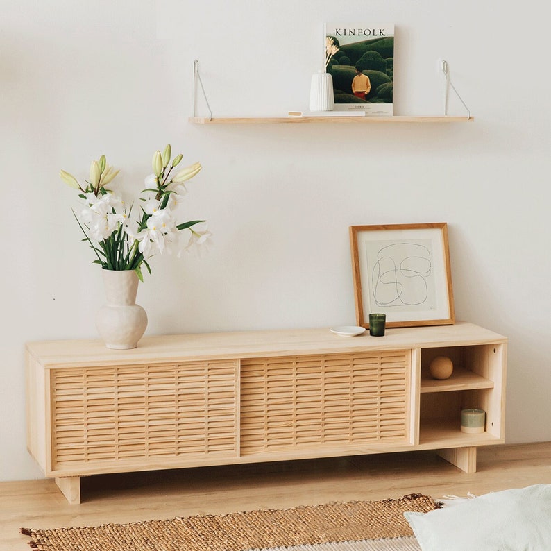 Puede incluir: Un armario de madera clara con dos puertas correderas y un estante. Las puertas tienen un patr&oacute;n tejido y el armario est&aacute; sobre un suelo de madera clara. Hay un jarr&oacute;n de flores blancas encima del armario y una impresi&oacute;n enmarcada en la pared detr&aacute;s.