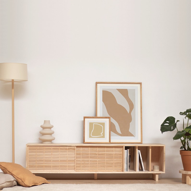Pu&ograve; includere: Un mobile TV in legno chiaro con due ante scorrevoli e ripiani aperti. Il mobile ha una finitura in legno chiaro e un design a doghe. Ci sono due stampe astratte incorniciate sul muro sopra il mobile, una pi&ugrave; grande dell'altra. Una pianta in vaso si trova a destra del mobile.