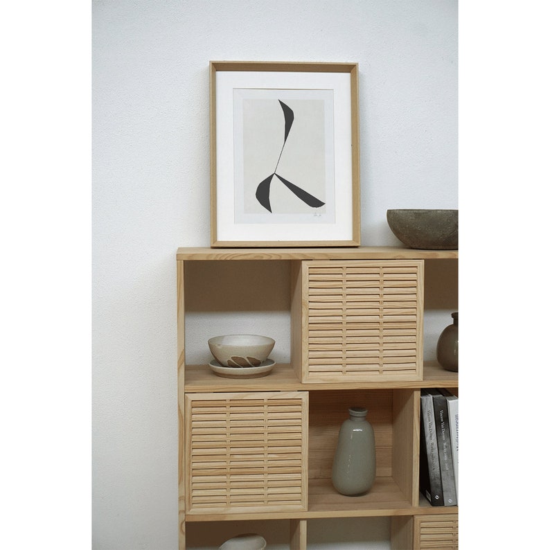 Op de afbeelding: Een houten rek met lamellen deuren en open planken. De planken zijn versierd met een ingelijst drukwerk, een kom, een vaas en boeken.