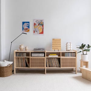 May include: A light wood record player stand with six shelves, two of which have slatted doors. The stand has a record player, amplifier, and a calendar on top. There are two vases and a white lamp on the top shelf. The stand is in front of a white wall with two framed pictures.