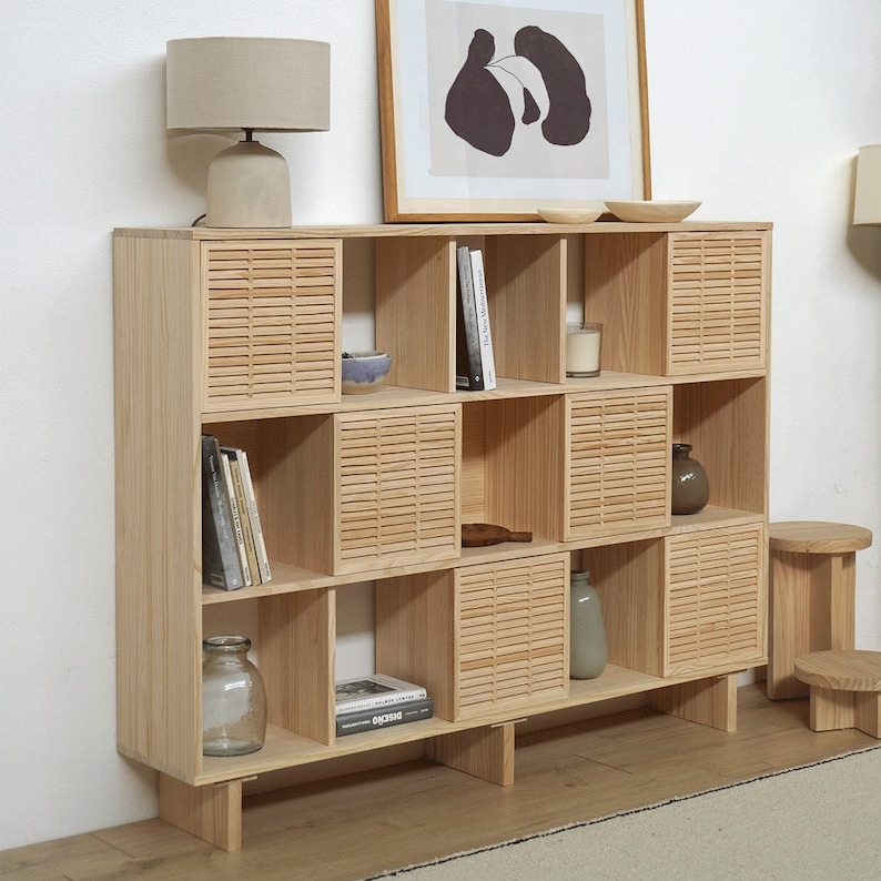 May include: A light wood bookshelf with six shelves and a slatted design. The shelves are filled with books, decorative items, and a candle. The bookshelf is standing on a light wood floor.