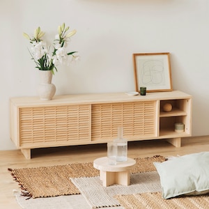Puede incluir: Un mueble de TV de madera clara con puertas correderas y estantes abiertos. El mueble tiene un acabado de madera natural y est&aacute; decorado con un jarr&oacute;n de flores blancas, una impresi&oacute;n enmarcada y dos candelabros de vidrio.