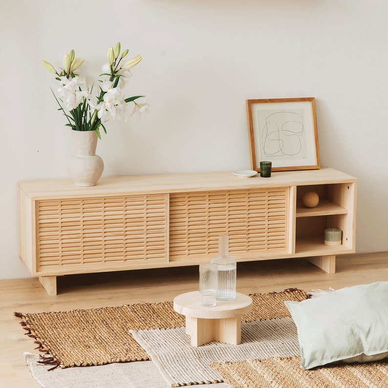 Puede incluir: Un mueble de TV de madera clara con puertas correderas y estantes abiertos. El mueble tiene un acabado de madera natural y est&aacute; decorado con un jarr&oacute;n de flores blancas, una impresi&oacute;n enmarcada y dos candelabros de vidrio.