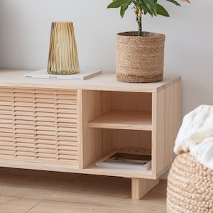Puede incluir: Un armario de madera clara con dos puertas y un estante. El armario tiene un dise&ntilde;o de listones en las puertas delanteras. El armario est&aacute; sobre un suelo de madera. Hay una planta en una cesta tejida encima del armario.