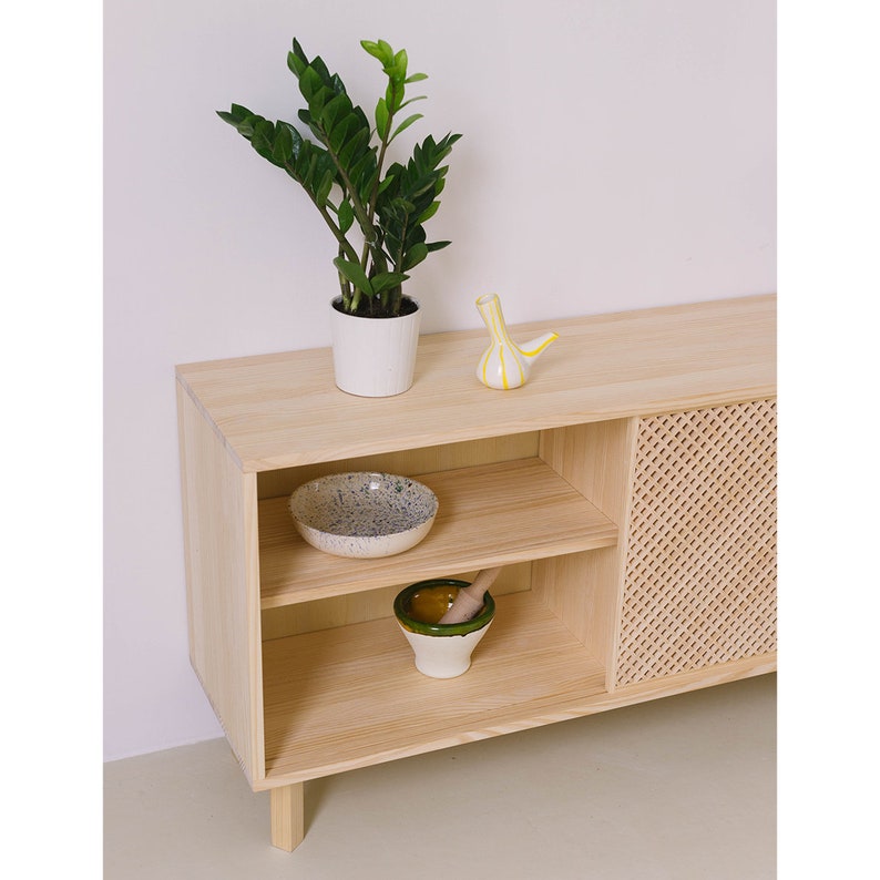 May include: A light wood cabinet with two shelves and a woven door. The cabinet has a plant in a white pot, a small yellow ceramic vase, a blue and white bowl, and a green mortar and pestle on the shelves.