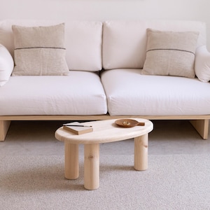 May include: A light wood oval coffee table with three cylindrical legs. The table is in front of a white sofa with two beige cushions. A small wooden tray and a notebook are on the table.