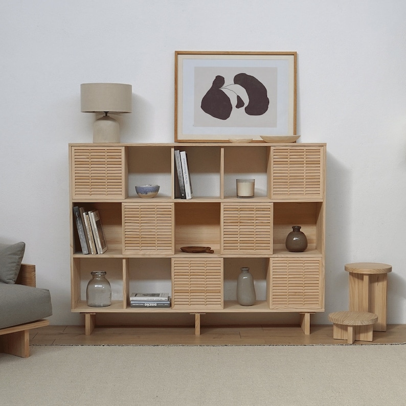 May include: A light wood bookshelf with 9 cubbies, each with a slatted front. The shelves are filled with books, decorative items, and a lamp. The bookshelf is on a white wall with a framed print above it.