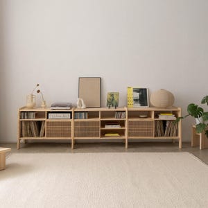 May include: A light wood entertainment unit with three sections, each with open shelves and slatted doors. The unit is filled with books, records, and decorative items. A framed picture and a lamp are on top of the unit.