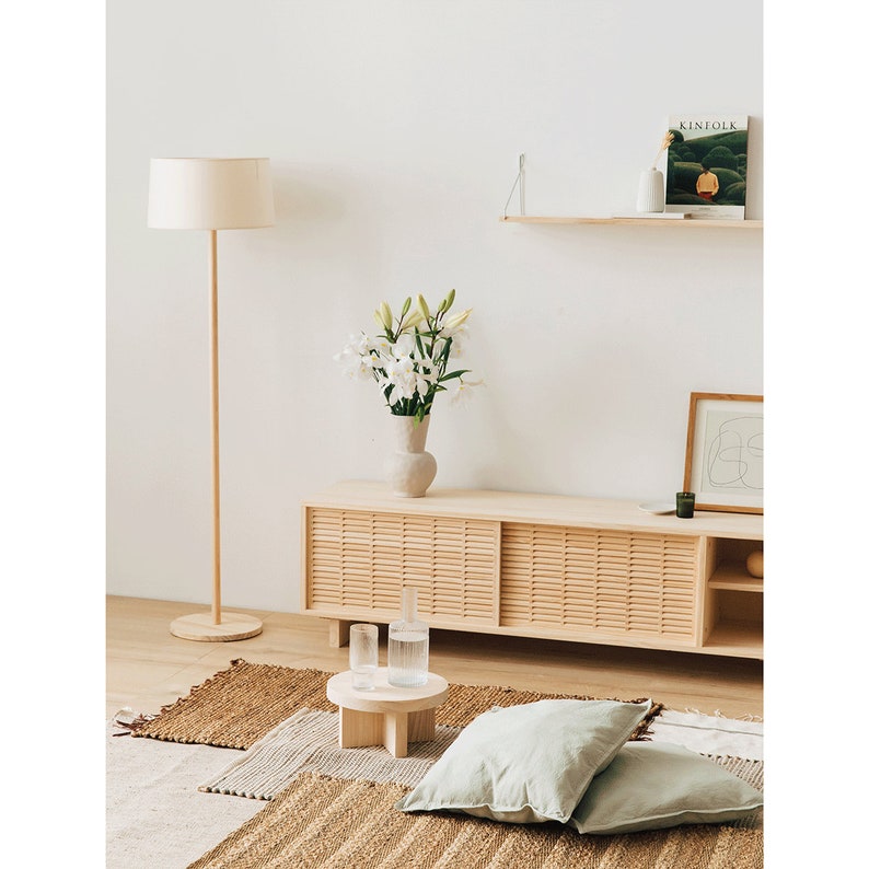 Puede incluir: Un centro de entretenimiento de madera clara con puertas corredizas y estantes abiertos. El centro est&aacute; decorado con un jarr&oacute;n de lirios blancos, una impresi&oacute;n enmarcada y una revista en un estante arriba. Una l&aacute;mpara de pie con una pantalla blanca se encuentra a la izquierda del centro de entretenimiento. Una alfombra tejida con una almohada de tiro verde claro se encuentra frente al centro de entretenimiento.
