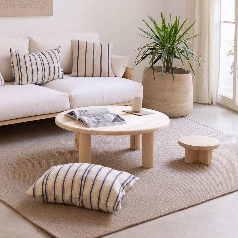 May include: A living room scene featuring a white sofa with striped pillows, a round wooden coffee table, and a small side table. A floor cushion with matching stripes and a large potted plant add to the room's decor. The rug is a neutral beige color.