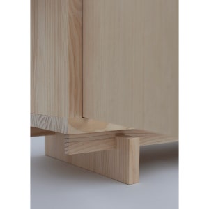 May include: A light wood cabinet with a single door and a simple design. The cabinet is raised on four rectangular wooden legs.