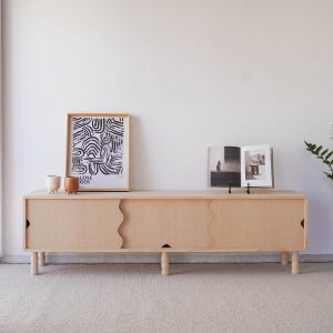 May include: A light wood cabinet with two sliding doors and four legs. The cabinet has a wavy design on the doors. There is a black and white print and two small ceramic pots on top of the cabinet.