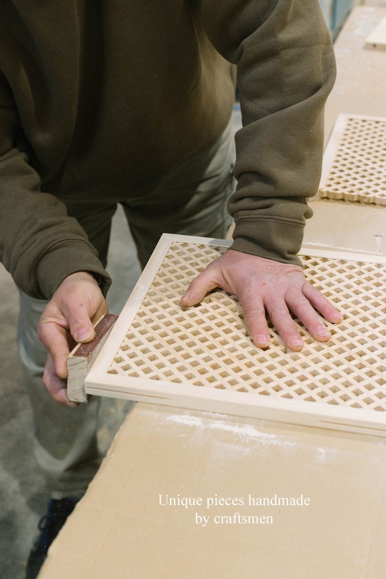 May include: A person's hand rests on a wooden lattice panel, which is being sanded by another hand holding a sanding block. The panel is made of light wood and has a grid pattern. The text "Unique pieces handmade by craftsmen" is visible at the bottom of the image.