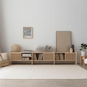 May include: A light wood record player stand with a slatted design. The stand has three sections, each with a shelf for records. There is a small black stereo receiver on the top shelf. The stand is in front of a white wall with a black and white framed print and a large framed canvas.