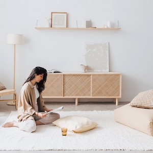 Puede incluir: Una mujer se sienta en una alfombra de pelo blanco en una habitaci&oacute;n luminosa y aireada, leyendo una revista. Lleva una camisa y pantalones beige. La habitaci&oacute;n tiene un armario de madera con un frente de rat&aacute;n tejido, una estanter&iacute;a con obras de arte enmarcadas y una l&aacute;mpara de pie.