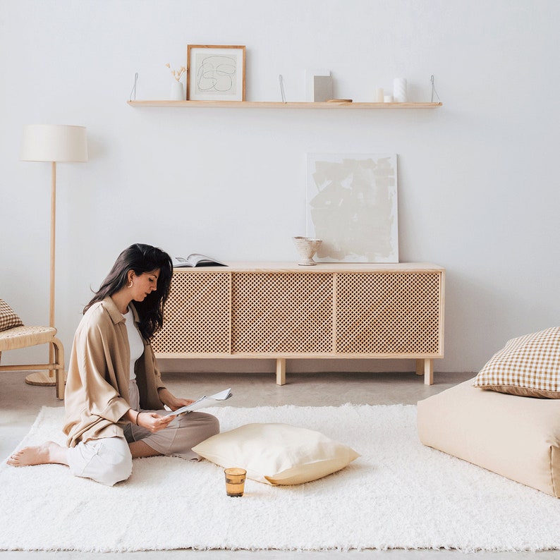 Puede incluir: Una mujer se sienta en una alfombra de pelo blanco en una habitaci&oacute;n luminosa y aireada, leyendo una revista. Lleva una camisa y pantalones beige. La habitaci&oacute;n tiene un armario de madera con un frente de rat&aacute;n tejido, una estanter&iacute;a con obras de arte enmarcadas y una l&aacute;mpara de pie.