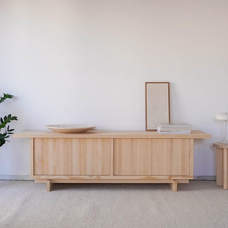 May include: A light wood cabinet with two sliding doors. The cabinet has a simple, minimalist design and is sitting on a light beige rug. There is a framed picture and a stack of books on top of the cabinet.