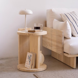 Può includere: Un comodino in legno chiaro con piano e base rotondi, con un ripiano integrato. Una lampada bianca, una tazza di vetro con un liquido scuro e un libro sono appoggiati sul tavolo. Un letto con cuscini bianchi è sullo sfondo.