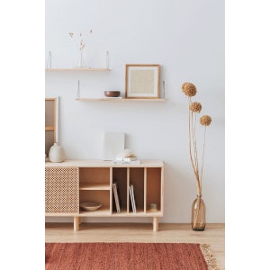 May include: A light wood cabinet with a woven door, open shelves, and a brown bowl on the top shelf. The cabinet is against a white wall with two floating shelves, a vase with dried flowers, and a framed picture.