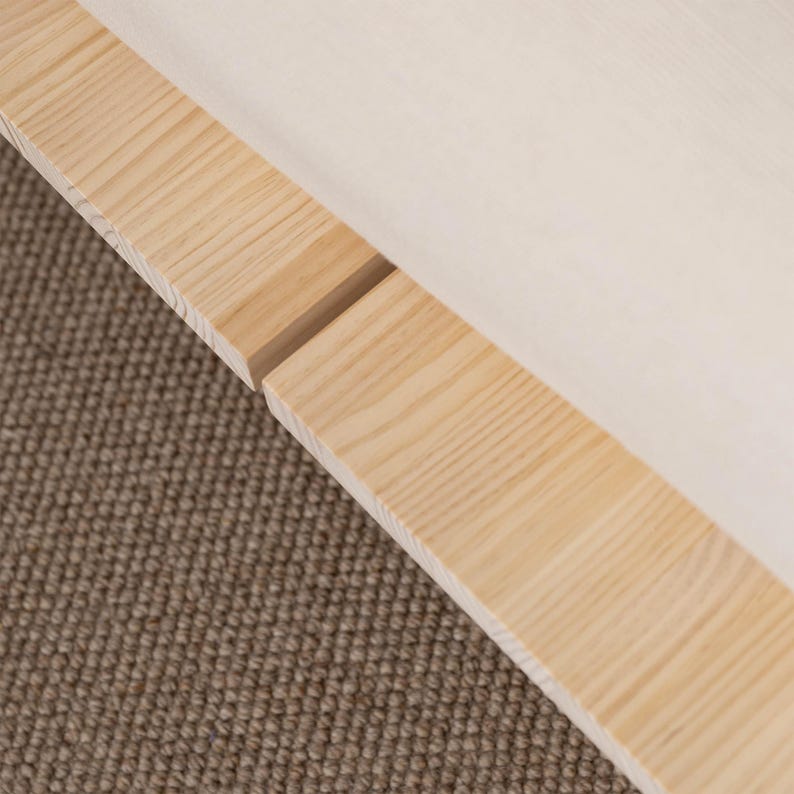 Puede incluir: Primer plano de un marco de madera de color claro con un acabado natural. El marco est&aacute; hecho de dos tablas de madera paralelas con un espacio entre ellas. Un tejido de color claro es visible en el fondo, descansando sobre una alfombra marr&oacute;n.