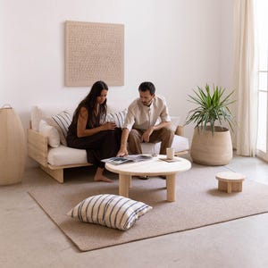 May include: A living room scene featuring a light wood sofa with white cushions and striped pillows. A round wooden coffee table and a small side table sit on a neutral rug. A large potted plant and decorative lamp are also present.