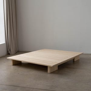 Puede incluir: Un somier de plataforma de madera de color claro. El somier es rectangular con un perfil bajo, sostenido por cuatro patas rectangulares. La madera tiene un acabado natural y el somier est&aacute; colocado sobre un suelo de hormig&oacute;n.