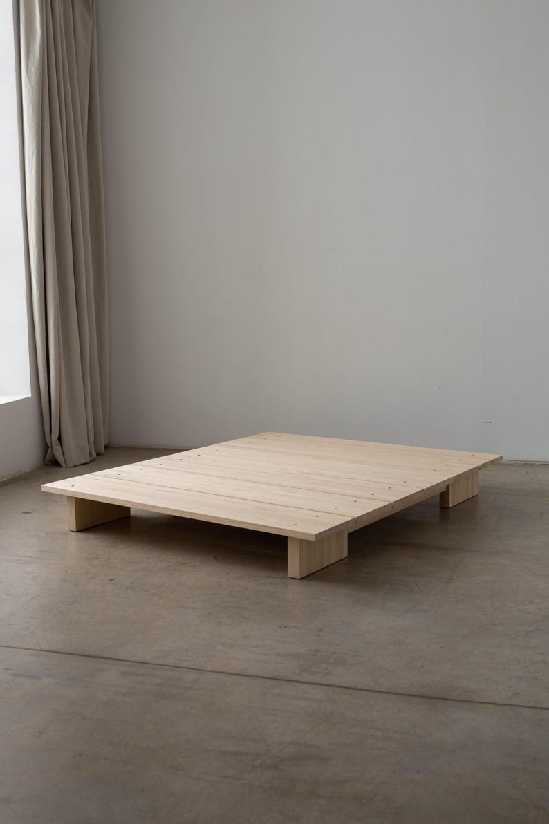 Puede incluir: Un somier de plataforma de madera de color claro. El somier es rectangular con un perfil bajo, sostenido por cuatro patas rectangulares. La madera tiene un acabado natural y el somier est&aacute; colocado sobre un suelo de hormig&oacute;n.