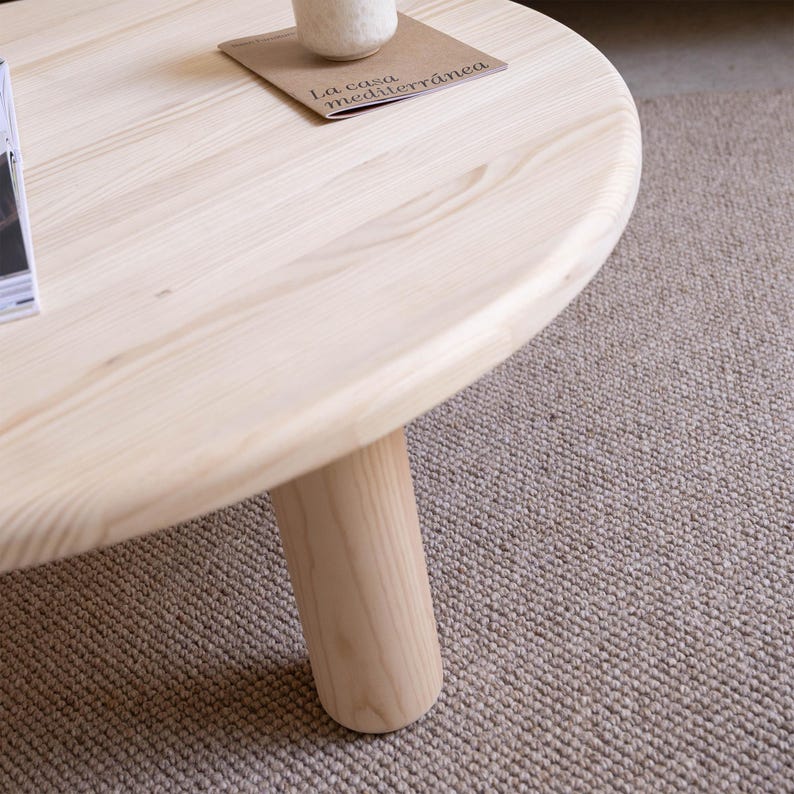 May include: A light-colored, round wooden coffee table with a single, cylindrical leg. The tabletop has a smooth finish. A small, rectangular card with text and a white ceramic object are on the table. The table sits on a neutral-colored rug.